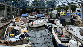 Boats docked at a busy marina with various equipment and reflections under a blue metal canopy.